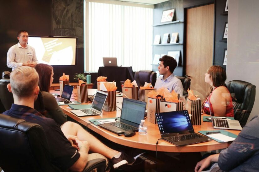 A marketing team with 5 members meeting around a table with one man making a presentation.