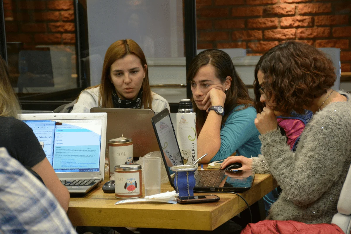 Four women collaborate around laptops at a wooden table in a cozy, brick-walled setting. They appear focused and engaged, surrounded by drinks and snacks.