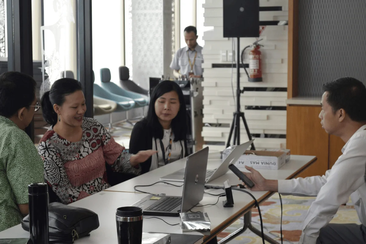 A group of four people in a modern office setting are engaged in a discussion around a table with laptops. One is talking animatedly, creating an atmosphere of collaboration.